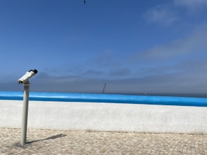 A telescope is positioned on a tiled promenade, facing the ocean. The horizon is visible under a mostly clear blue sky with some clouds. A seagull flies high above, and a distant crane or structure can be seen near the water.