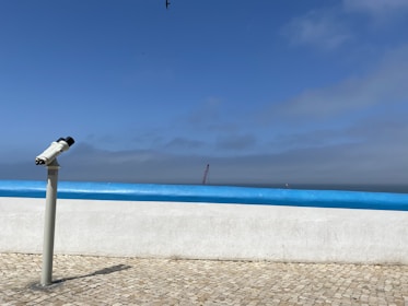 A telescope is positioned on a tiled promenade, facing the ocean. The horizon is visible under a mostly clear blue sky with some clouds. A seagull flies high above, and a distant crane or structure can be seen near the water.