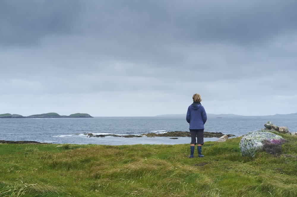 woman in black jacket standing on green grass field near sea during daytime