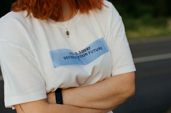 A person wearing a white t-shirt with a motivational message, 'YOUR TODAY DEFINES YOUR FUTURE', written on it. The person has red hair and is outdoors with arms crossed, wearing a black wristband and a pendant necklace.