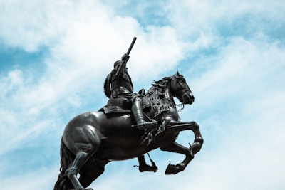 A statue of a warrior on horseback, depicted in a dynamic pose with the horse rearing up on its hind legs. The warrior is holding a weapon and wearing detailed armor. The statue is silhouetted against a backdrop of a partially cloudy sky.
