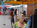 A bustling outdoor market scene with several people shopping and browsing various stalls. The market is covered with colorful canopies, and one stall features flowers, while another displays a selection of watches or jewelry. Some people are casually dressed, and there are both men and women visible. The overall environment is lively and crowded.
