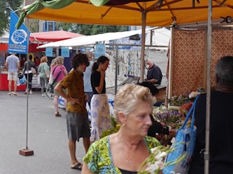 A bustling outdoor market scene with several people shopping and browsing various stalls. The market is covered with colorful canopies, and one stall features flowers, while another displays a selection of watches or jewelry. Some people are casually dressed, and there are both men and women visible. The overall environment is lively and crowded.