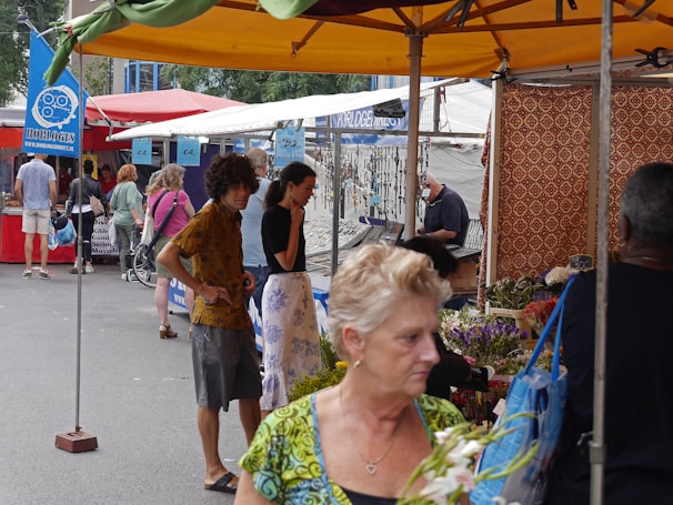 A bustling outdoor market scene with several people shopping and browsing various stalls. The market is covered with colorful canopies, and one stall features flowers, while another displays a selection of watches or jewelry. Some people are casually dressed, and there are both men and women visible. The overall environment is lively and crowded.