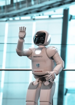 A humanoid robot is raising its hand in a sleek, modern environment. The robot, predominantly white with some branding, features a smooth design and a visor-like head.