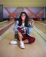 A person is sitting on a bowling alley lane, blowing a pink bubble with gum. The individual is wearing a denim jacket, a graphic T-shirt, red pants, and white sneakers. The bowling pins are arranged in the background with colorful lighting adding to the ambiance.