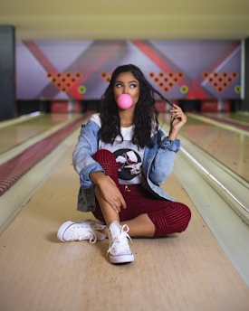 A person is sitting on a bowling alley lane, blowing a pink bubble with gum. The individual is wearing a denim jacket, a graphic T-shirt, red pants, and white sneakers. The bowling pins are arranged in the background with colorful lighting adding to the ambiance.