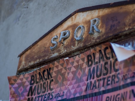 A weathered, rusty metal sign with the letters 'SPQR' embossed in a faded blue color, juxtaposed with a torn poster below that reads 'BLACK MUSIC MATTERS VOL. 2' in bold black print over a patterned background.