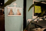 A framed religious artwork depicting two figures under a cross is mounted on a textured, light blue wall. To the right, there is a potted plant with elongated leaves. A bottle is seen on top of a table, next to some plastic chairs. A bright yellow sign with partially visible text is in the background.
