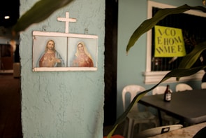 A framed religious artwork depicting two figures under a cross is mounted on a textured, light blue wall. To the right, there is a potted plant with elongated leaves. A bottle is seen on top of a table, next to some plastic chairs. A bright yellow sign with partially visible text is in the background.