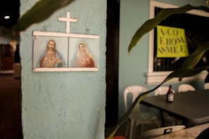 A framed religious artwork depicting two figures under a cross is mounted on a textured, light blue wall. To the right, there is a potted plant with elongated leaves. A bottle is seen on top of a table, next to some plastic chairs. A bright yellow sign with partially visible text is in the background.