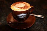 A brown ceramic cup filled with frothy cappuccino topped with a dusting of cocoa powder sits on a matching saucer. A small spoon rests on the saucer's edge. The cup has a logo that reads 'Caffe Italia Dal 1954.' The background is a dark surface, creating a warm and inviting atmosphere.