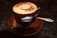 A brown ceramic cup filled with frothy cappuccino topped with a dusting of cocoa powder sits on a matching saucer. A small spoon rests on the saucer's edge. The cup has a logo that reads 'Caffe Italia Dal 1954.' The background is a dark surface, creating a warm and inviting atmosphere.
