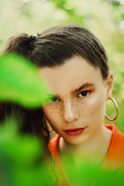 A woman with dark hair tied back is partially obscured by vibrant green leaves. Her face is prominently featured with bold freckles, striking eyebrows, and red lipstick. She is wearing a hoop earring and an orange top.