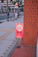 A sidewalk with a bright red sign advertising a used shop that sells CDs, DVDs, and records. The sign leans against a brick building. In the background, there are two bicycles parked by a pole, a pedestrian crossing sign, and a few trees lining the street. The street appears quiet with modern buildings across the road.