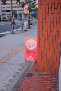 A sidewalk with a bright red sign advertising a used shop that sells CDs, DVDs, and records. The sign leans against a brick building. In the background, there are two bicycles parked by a pole, a pedestrian crossing sign, and a few trees lining the street. The street appears quiet with modern buildings across the road.