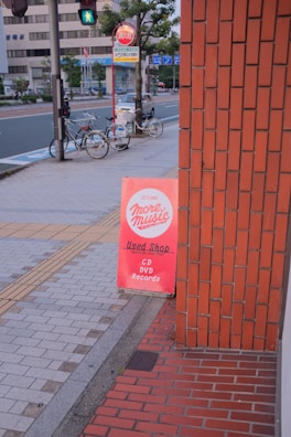 A sidewalk with a bright red sign advertising a used shop that sells CDs, DVDs, and records. The sign leans against a brick building. In the background, there are two bicycles parked by a pole, a pedestrian crossing sign, and a few trees lining the street. The street appears quiet with modern buildings across the road.