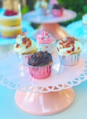 A group of assorted cupcakes presented on a decorative pink cake stand. The cupcakes are adorned with a variety of toppings including sprinkles, chocolate, and bacon bits. Each cupcake is wrapped in colorful foil, and the setting appears to be bright and airy, suggesting a celebratory or festive occasion.