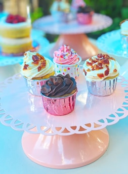 A group of assorted cupcakes presented on a decorative pink cake stand. The cupcakes are adorned with a variety of toppings including sprinkles, chocolate, and bacon bits. Each cupcake is wrapped in colorful foil, and the setting appears to be bright and airy, suggesting a celebratory or festive occasion.