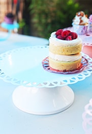 Two small layered cakes are adorned with a swirl of cream and topped with fresh raspberries. They are presented on a delicate, white, lace-patterned cake stand. The background features a slightly blurred, colorful array of cupcakes, adding to the lively and inviting ambiance.