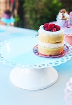 Two small layered cakes are adorned with a swirl of cream and topped with fresh raspberries. They are presented on a delicate, white, lace-patterned cake stand. The background features a slightly blurred, colorful array of cupcakes, adding to the lively and inviting ambiance.