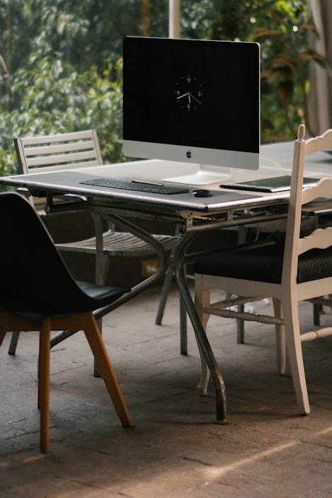 A desktop computer with a keyboard and mouse sits on a metal table in a room with several chairs around it. The computer screen displays a clock, and the setting is well-lit, surrounded by greenery visible through large windows.