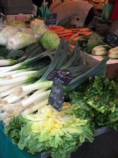 A vibrant market stall displays a variety of fresh vegetables. The assortment includes leeks, cabbages, carrots, and different types of leafy greens. Chalkboard signs with prices are visible, and the arrangement is abundant and colorful.