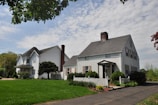 Two traditional-style houses are positioned along a quiet street, each with white siding and dark shingled roofs. The houses are surrounded by well-maintained lawns and landscaped gardens, featuring shrubs and small trees. A clear sky with scattered clouds forms the backdrop, adding to the peaceful suburban setting.