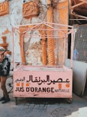 A weathered juice stand, painted in pink, displays Arabic and French text advertising orange juice. It stands in front of a textured wall with ornate carvings and orange columns. A partially visible bicycle and a person standing nearby can be seen.