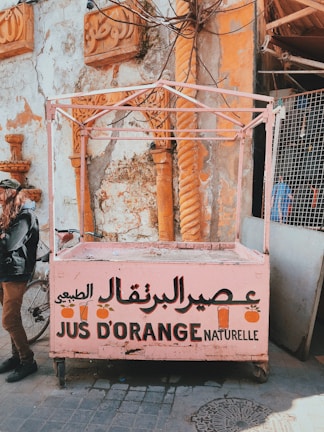 A weathered juice stand, painted in pink, displays Arabic and French text advertising orange juice. It stands in front of a textured wall with ornate carvings and orange columns. A partially visible bicycle and a person standing nearby can be seen.