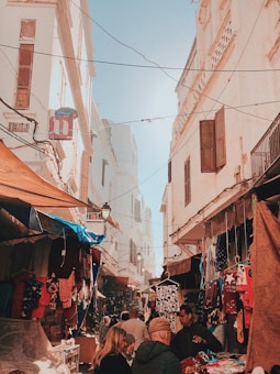 A bustling street market in a historical setting with narrow alleyways lined by whitewashed buildings. People are browsing and shopping for various colorful clothing items and textiles hung on display racks under makeshift stalls. The market is covered with fabric canopies, creating a vibrant and lively atmosphere.