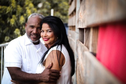 An older couple stands close together, smiling and embracing warmly. They are dressed in white clothing, with the background displaying a blurred mix of green foliage and a wooden structure.
