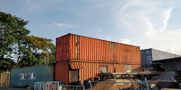Several shipping containers are stacked in an outdoor area, with some covered by tarps. The containers are mostly in shades of orange, blue, and gray. There are trees in the background, and the sky is mostly clear with a few clouds.