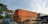 Several shipping containers are stacked in an outdoor area, with some covered by tarps. The containers are mostly in shades of orange, blue, and gray. There are trees in the background, and the sky is mostly clear with a few clouds.