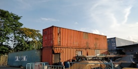 Several shipping containers are stacked in an outdoor area, with some covered by tarps. The containers are mostly in shades of orange, blue, and gray. There are trees in the background, and the sky is mostly clear with a few clouds.