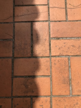 A section of red-orange floor tiles, each tile rectangular in shape, arranged in a regular pattern with visible grout lines in between. The tiles exhibit some signs of wear and darker patches, suggesting age or frequent use. A shadow is cast across part of the tiles, adding contrast to the composition.