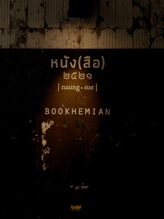 A dimly lit, textured wall with peeling paint serves as the background. There is Thai script accompanied by the phonetic pronunciation 'naung.sue' and the English word 'BOOKHEMIAN' illuminated by the light.