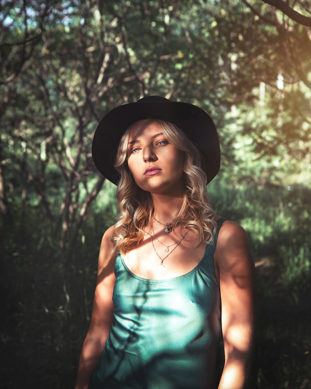 woman in blue spaghetti strap top wearing black fedora hat standing in forest during daytime