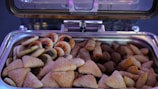 An open buffet tray is filled with a variety of small, savory pastries. The assortment includes sesame-topped triangular pastries, mini pizzas with visible toppings, spring rolls, kibbeh, and other fried or baked snacks.
