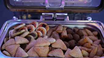 An open buffet tray is filled with a variety of small, savory pastries. The assortment includes sesame-topped triangular pastries, mini pizzas with visible toppings, spring rolls, kibbeh, and other fried or baked snacks.