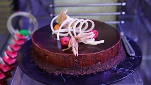 A beautifully crafted chocolate cake on a dark platter, adorned with white chocolate curls and garnished with raspberries and cape gooseberries. The cake has a smooth, glossy finish and is surrounded by a layer of chocolate crumbs.