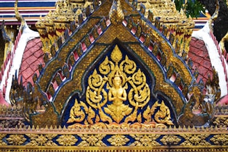 Intricate golden carvings and ornate patterns adorn a traditional architectural structure, featuring a central figure in a meditative pose surrounded by floral motifs. The detailed craftsmanship showcases vibrant colors and elaborate designs typical of Southeast Asian temple architecture.