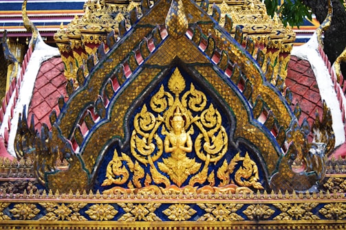 Intricate golden carvings and ornate patterns adorn a traditional architectural structure, featuring a central figure in a meditative pose surrounded by floral motifs. The detailed craftsmanship showcases vibrant colors and elaborate designs typical of Southeast Asian temple architecture.