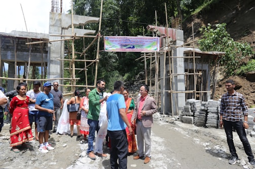 A group of people are gathered near a construction site with partially built structures and scaffolding. Some individuals are holding bags, and there is a banner overhead. The site is surrounded by greenery and building materials like bricks are stacked on the side.