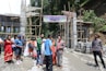 A group of people are gathered near a construction site with partially built structures and scaffolding. Some individuals are holding bags, and there is a banner overhead. The site is surrounded by greenery and building materials like bricks are stacked on the side.