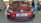 A red Volkswagen car with TSI Power branding on the hood is parked inside a garage. The front bumper has decals and a license plate featuring 'Venky's xprs'. Two people are visible in the background, possibly mechanics, standing near a workbench and another car.