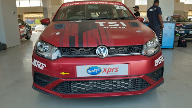 A red Volkswagen car with TSI Power branding on the hood is parked inside a garage. The front bumper has decals and a license plate featuring 'Venky's xprs'. Two people are visible in the background, possibly mechanics, standing near a workbench and another car.