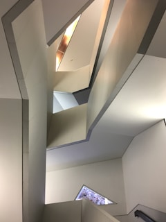 A geometrical architectural space with angular walls, varying shades of gray and beige. The design features multiple reflective surfaces and sharp lines creating an abstract pattern. Natural light filters through narrow openings, adding depth and highlighting the minimalist aesthetic.