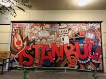 A vibrant mural features the word 'Istanbul' in bold, red letters, surrounded by artistic depictions of iconic city landmarks and elements including domes, towers, and traditional symbols. The mural is colorful and displays a creative urban vibe. There is a wooden bench to the left of the mural and some leafy branches hanging down from the upper left corner.