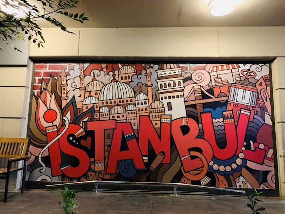 A vibrant mural features the word 'Istanbul' in bold, red letters, surrounded by artistic depictions of iconic city landmarks and elements including domes, towers, and traditional symbols. The mural is colorful and displays a creative urban vibe. There is a wooden bench to the left of the mural and some leafy branches hanging down from the upper left corner.